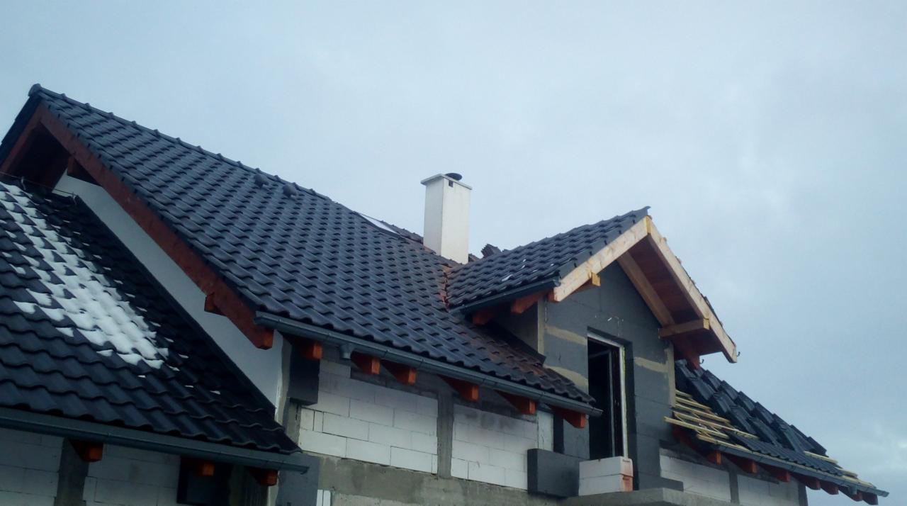 Częściowo pokryty ciemną dachówką dach domu w trakcie budowy, widoczny komin, część dachu pokryta śniegiem, elementy drewniane konstrukcji dachowej.