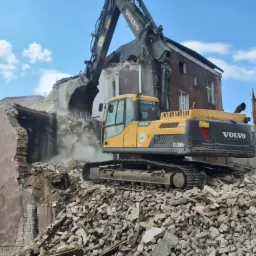 Żółta koparka Volvo EC250D na gruzowisku podczas rozbiórki budynku, w tle budynek i wieża kościoła pod błękitnym niebem.