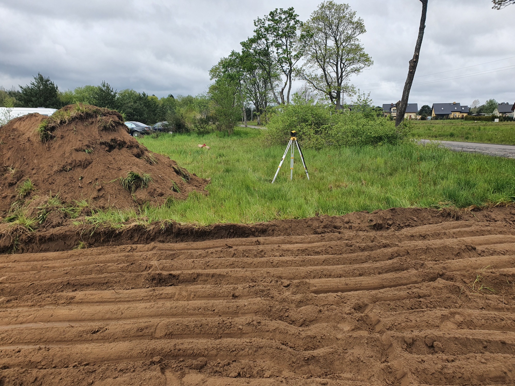Świeżo wyrównany teren z widocznymi śladami po maszynach, pryzma ziemi z darnią i niwelator laserowy na trójnogu w tle, krajobraz wiejski.