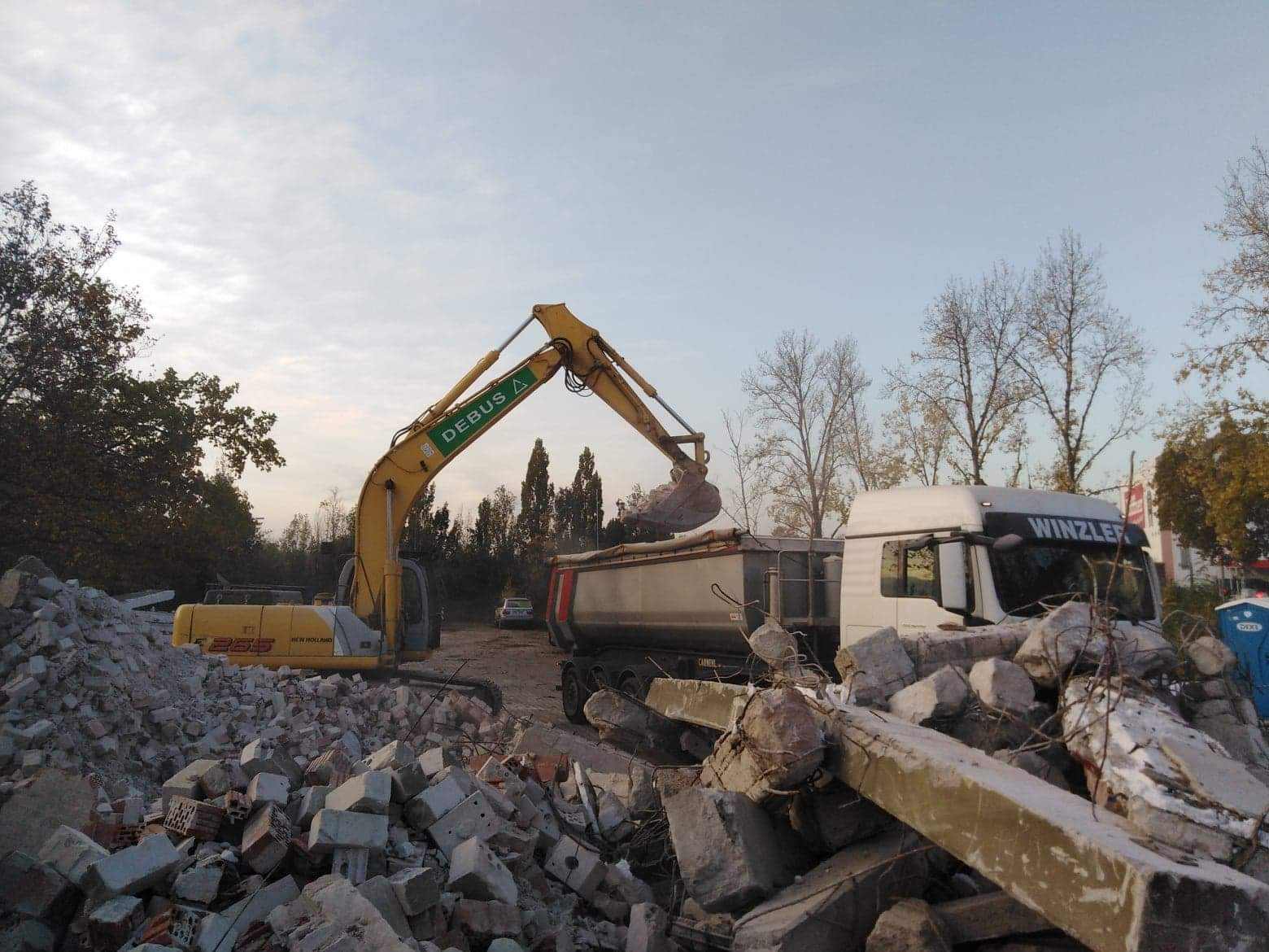 Żółta koparka DEBUS załadowuje gruz do skrzyni ciężarówki WINZLER na tle drzew i bladego nieba, z hałdą gruzu na pierwszym planie.