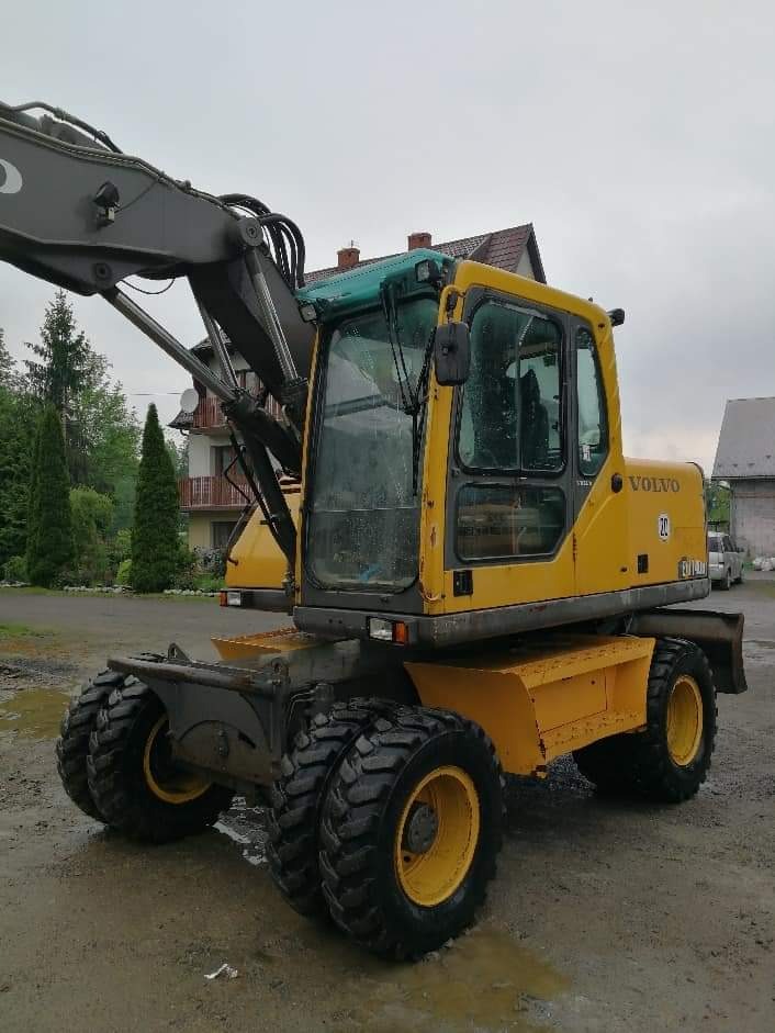 Żółta koparka kołowa Volvo w błotnistym terenie, z widocznymi budynkami mieszkalnymi w tle i pochmurnym niebem.