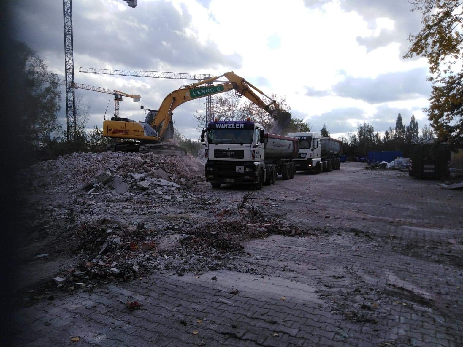 Żółta koparka z długim ramieniem załadowuje gruz na białą ciężarówkę MAN Winzler, w tle drugi podobny pojazd i dźwigi budowlane.