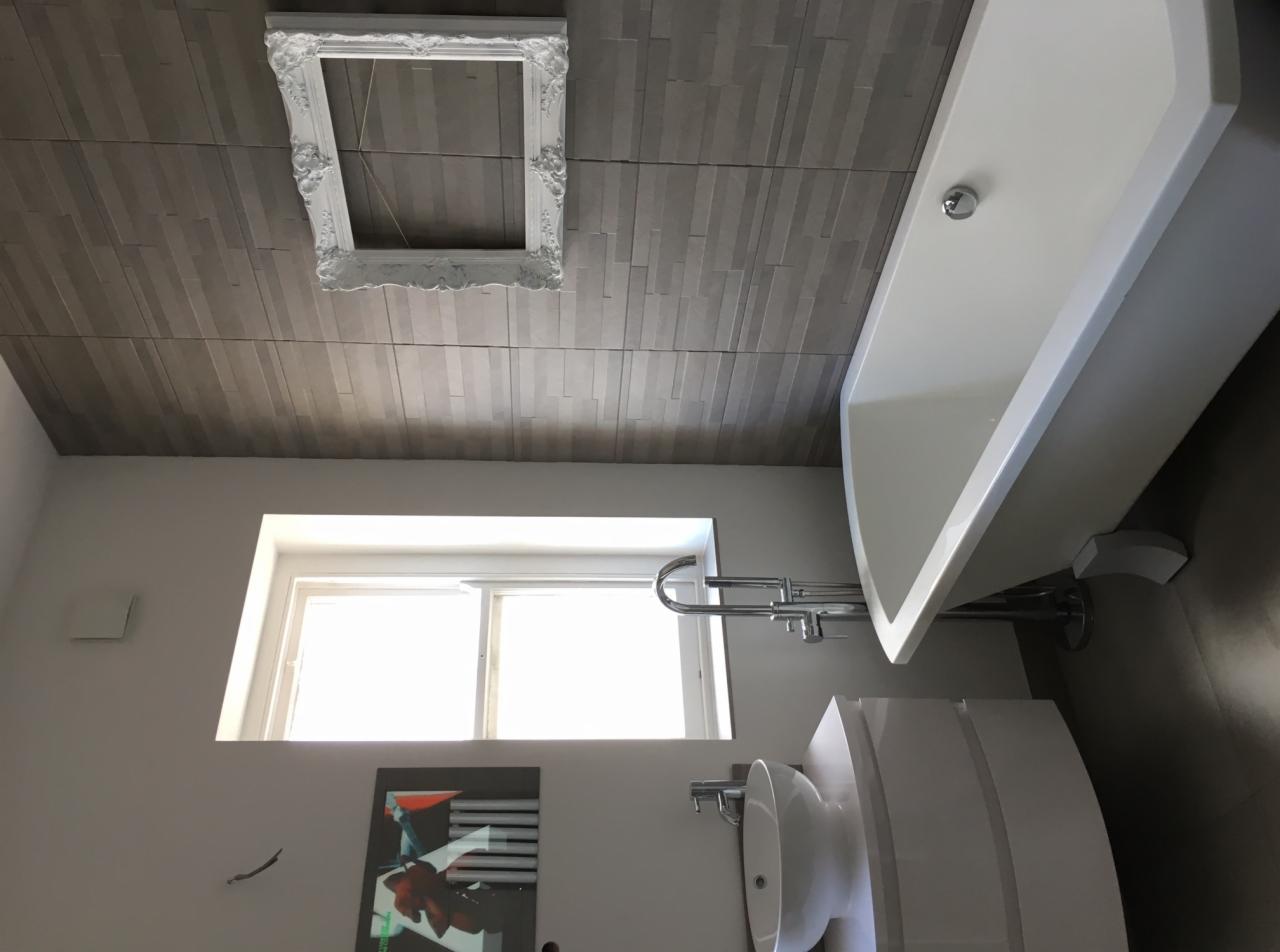 Nowoczesna łazienka z białą wolnostojącą wanną, umywalką na szafce o zaokrąglonym kształcie, szarą okładziną ścienną imitującą drewno i dekoracyjną ramą na suficie.