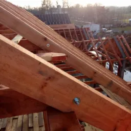 Konstrukcja drewniana dachu w trakcie budowy, widoczne elementy mocujące, w tle budynki i drzewa.