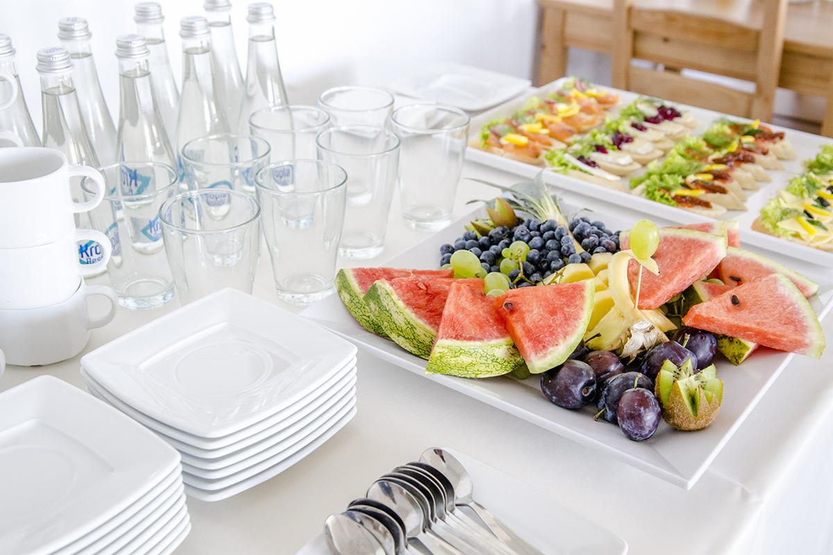 Stół z białym obrusem zastawiony talerzami, szklankami, butelkami wody i dwoma półmiskami: jeden z przekąskami, drugi z owocami (arbuz, śliwki, winogrona, kiwi) i ananasem.