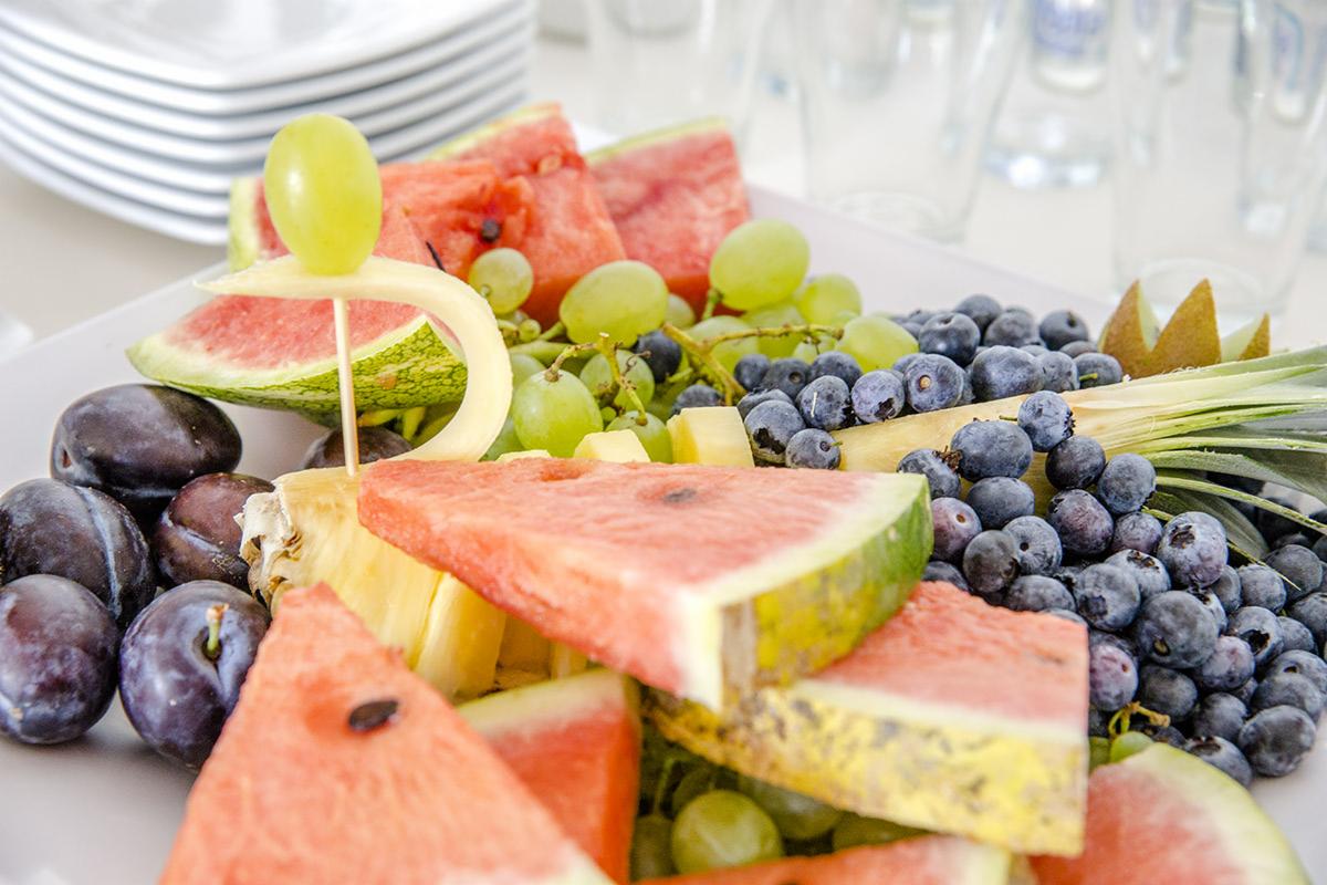 Dekoracyjna kompozycja z owoców: plastry arbuza, winogrona, śliwki, borówki i ananas na białym talerzu, z wykałaczką i spiralnie obranym kawałkiem melona jako ozdobą, w tle stos białych talerzy...