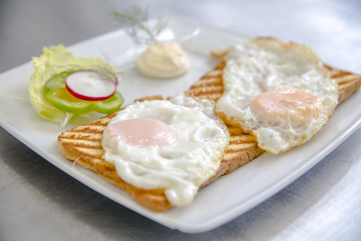 Dwa jajka sadzone z płynnym żółtkiem na grzankach, podane z sałatką z sałaty, papryki i rzodkiewki oraz sosem na białym, kwadratowym talerzu.