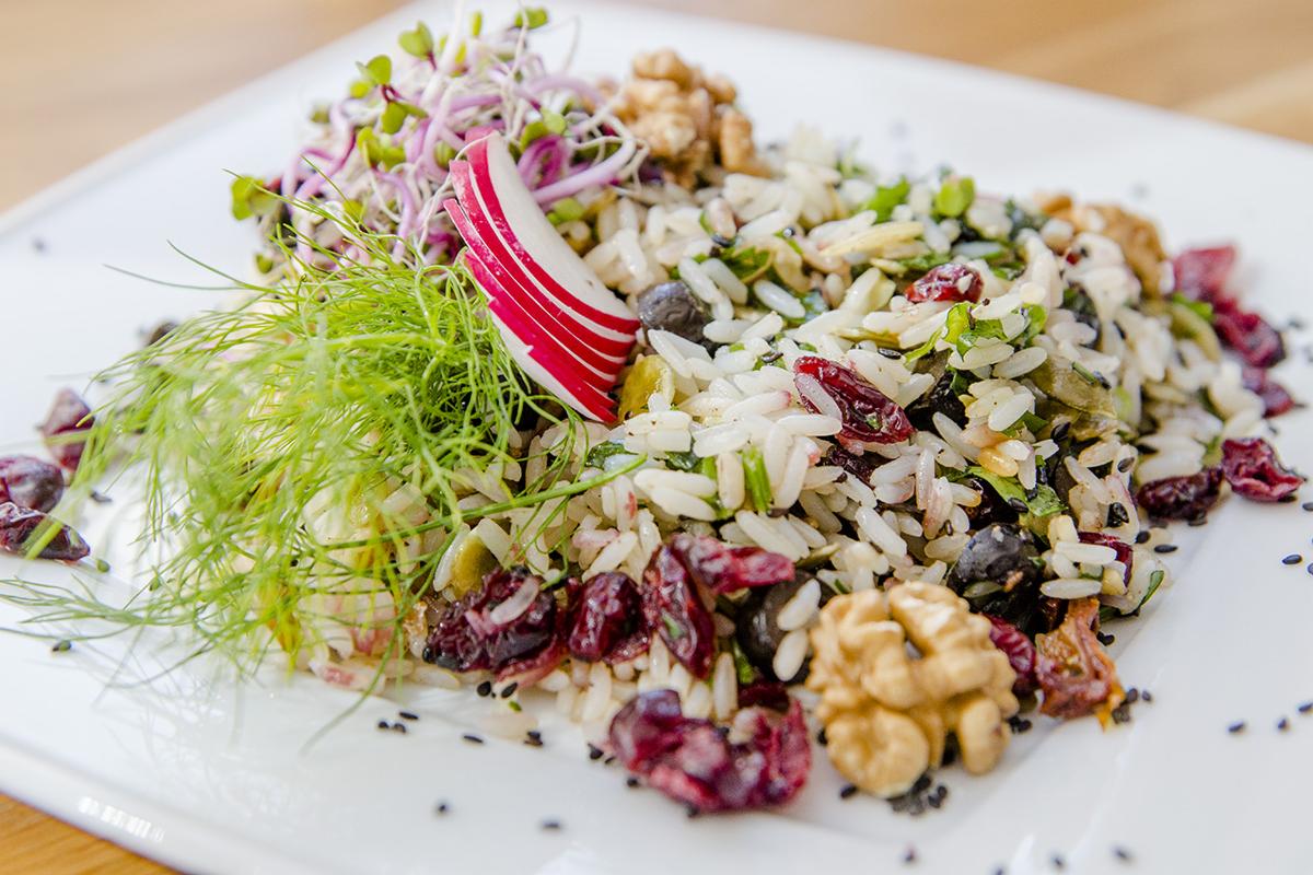 Ułożona na białym kwadratowym talerzu porcja sałatki ryżowej z żurawiną, orzechami włoskimi, rzodkiewką w paski, kiełkami i koperkiem włoskim, posypana ziarnami czarnego sezamu.