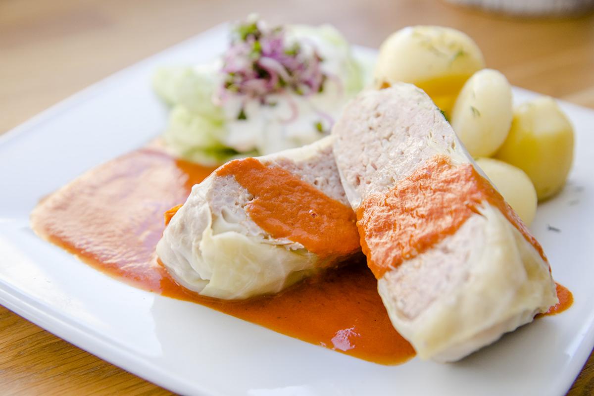 Dwa przekrojone gołąbki polane pomarańczowym sosem na białym talerzu, podane z młodymi ziemniakami i surówką z sosem śmietanowym.