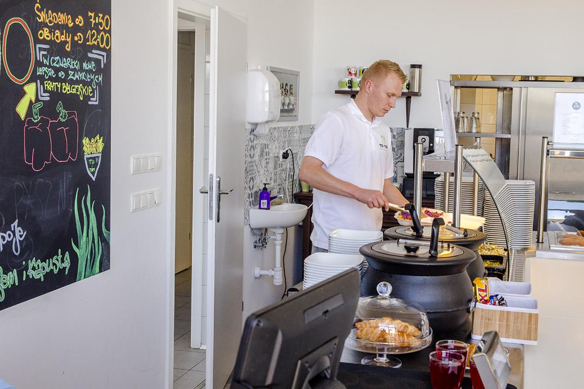 Pracownik w białej koszulce nakłada sałatkę na talerz z dużego, czarnego garnka w punkcie gastronomicznym z tablicą menu wypisaną kredą w tle.