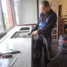 Pracownik w niebieskim kombinezonie szlifuje granitowy blat przy oknie za pomocą szlifierki kątowej podłączonej do odkurzacza przemysłowego, na blacie leżą narzędzia.