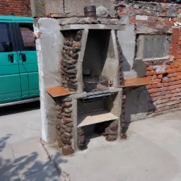 Murowany grill ogrodowy z kamienną obudową, częściowo otynkowany, z elementami z płytek ceramicznych, na betonowym podłożu, obok widoczny fragment zielonego samochodu dostawczego i ceglany budynek...