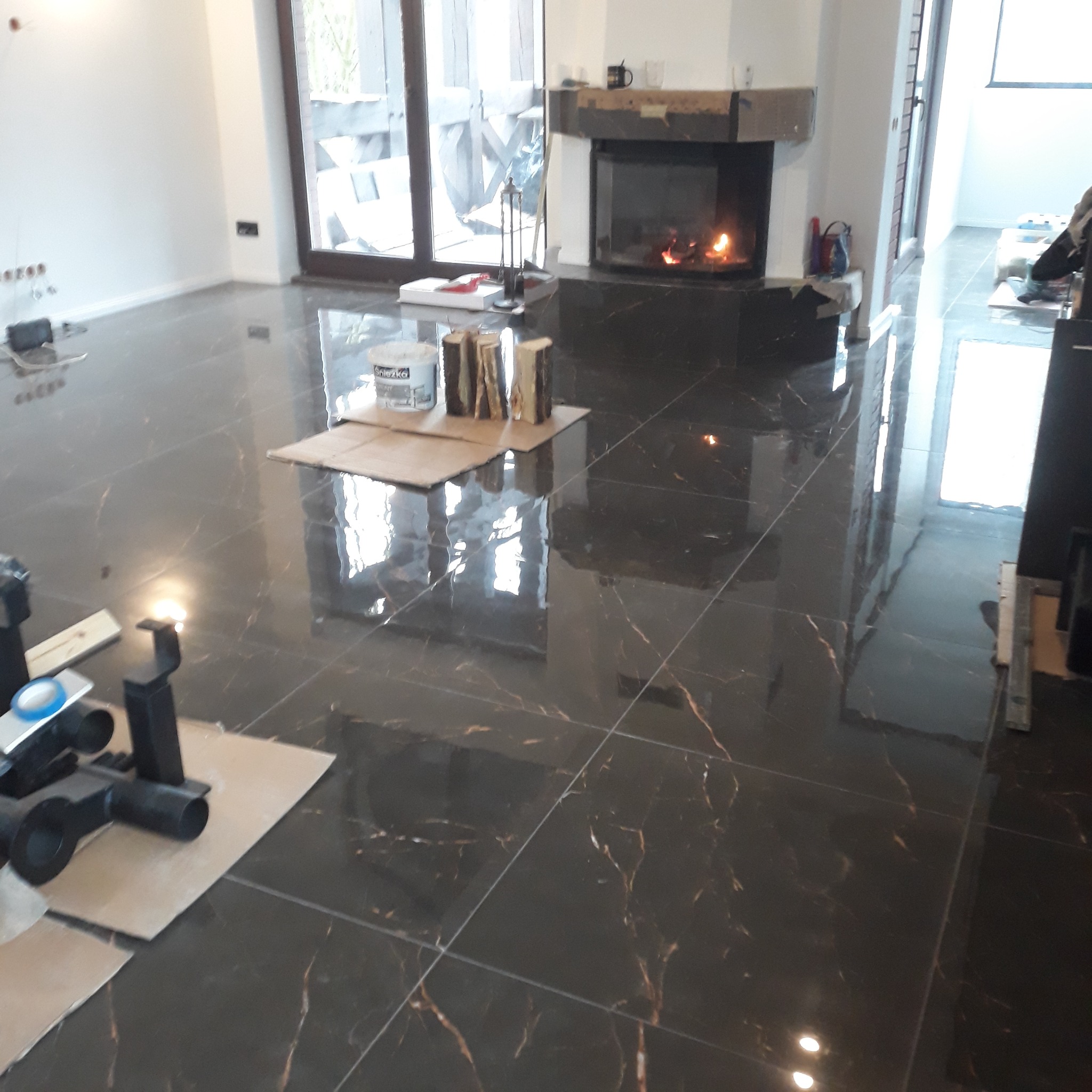 Położona, błyszcząca, ciemna podłoga z imitacją marmuru, widoczny kominek w tle, przygotowane rury kanalizacyjne i wiadro z zaprawą na kartonach.