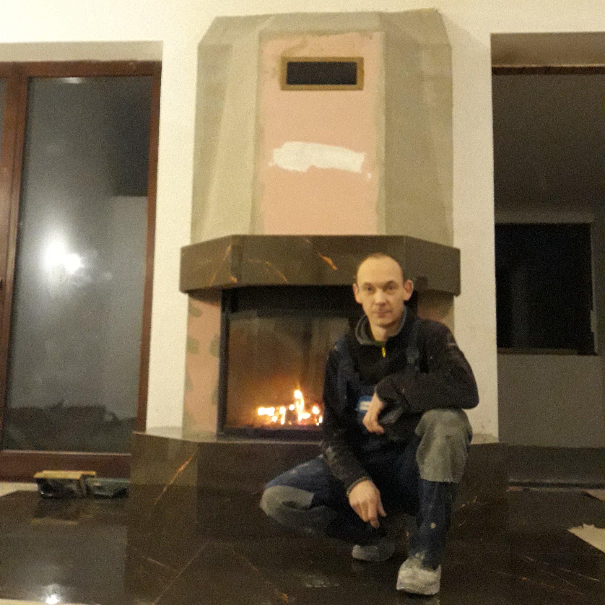 Mężczyzna w roboczym ubraniu kuca na podłodze z ciemnego marmuru przed nowo wykończonym kominkiem z marmurową obudową, w kominku pali się ogień, widoczne ślady po pracach wykończeniowych na ścianie.
