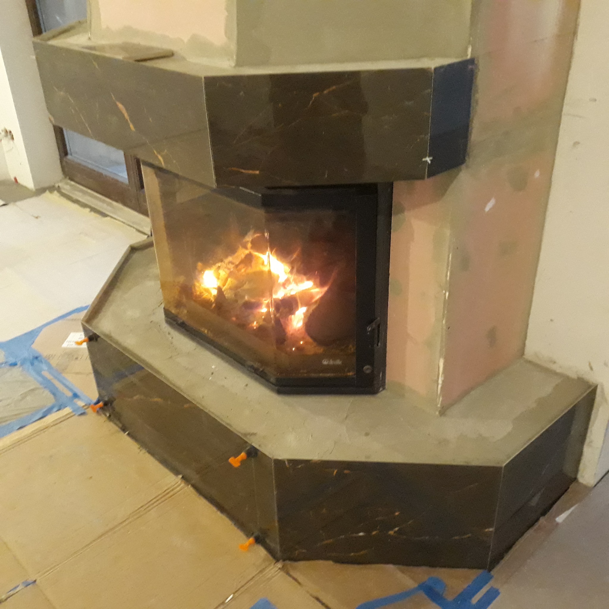 Obudowa kominka wykończona płytkami imitującymi ciemny marmur z widocznym paleniskiem i płomieniami, na podłodze karton zabezpieczony folią i krzyżyki dystansowe do płytek.