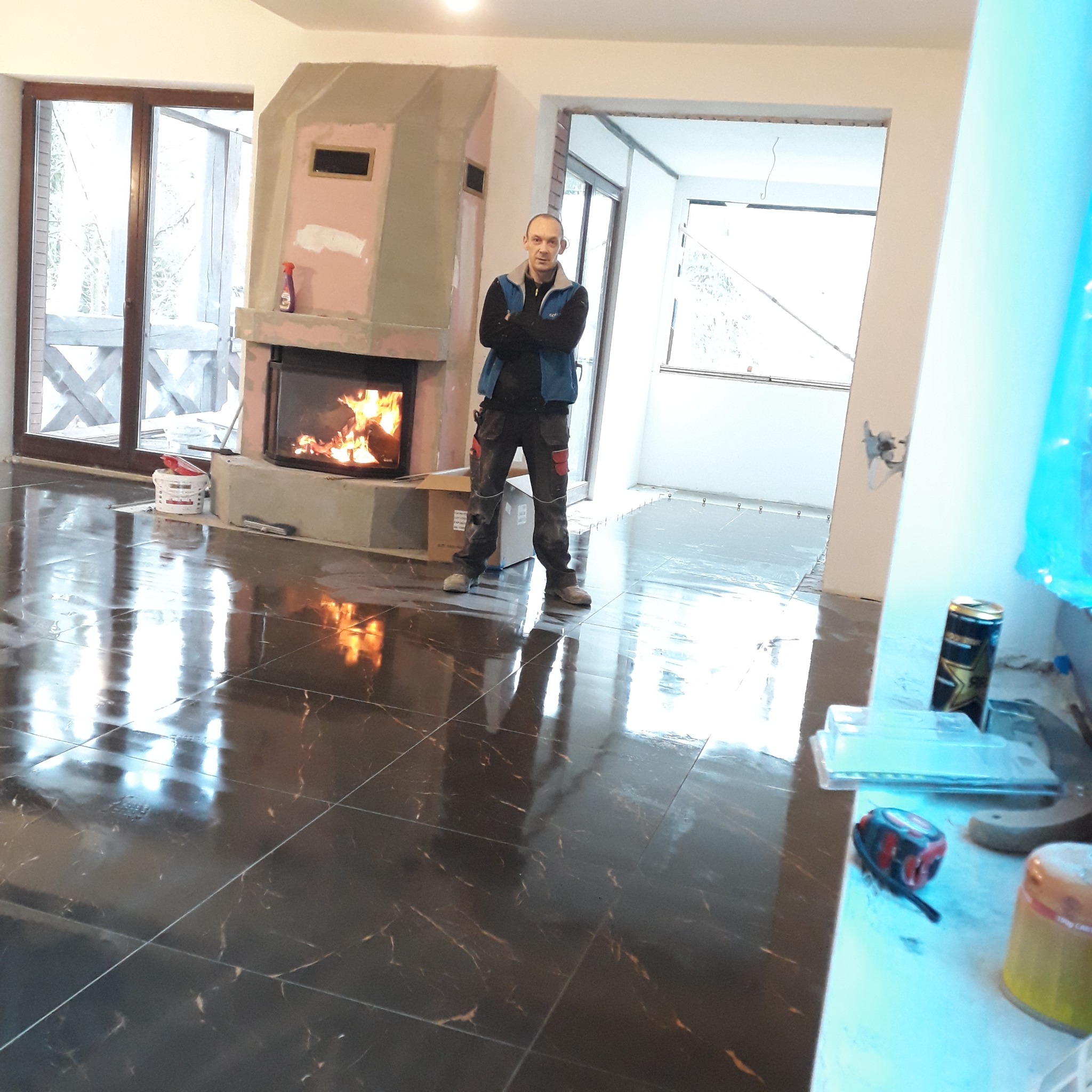 Układanie ciemnych, błyszczących płytek podłogowych z marmurowym wzorem w przestronnym salonie z kominkiem; pracownik w roboczym stroju stoi na ułożonej posadzce.