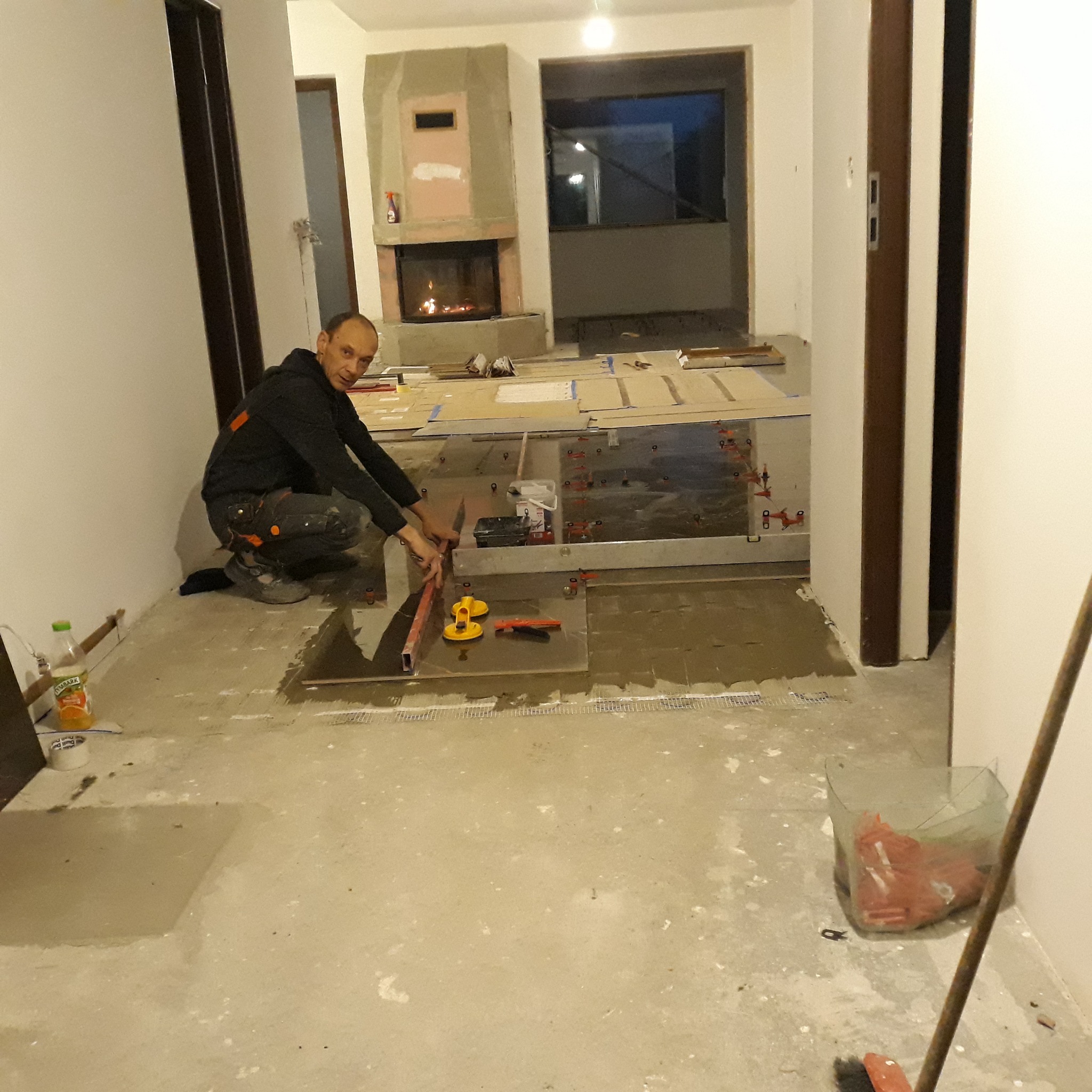 Mężczyzna w trakcie układania płytek podłogowych w pomieszczeniu z kominkiem, używa poziomicy i przyssawek do precyzyjnego ułożenia płytek.