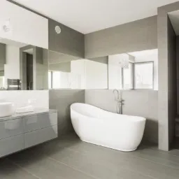 Nowoczesna łazienka w szarościach z białą, wolnostojącą wanną, umywalką na szafce z szufladami i lustrami na ścianie.