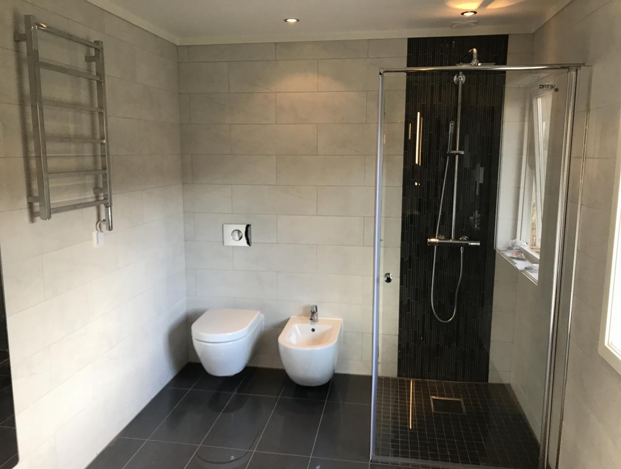 Nowoczesna łazienka z białymi płytkami imitującymi kamień, podwieszaną toaletą i bidetem, kabiną prysznicową ze szklanymi drzwiami i czarną mozaiką oraz grzejnikiem drabinkowym na ścianie.