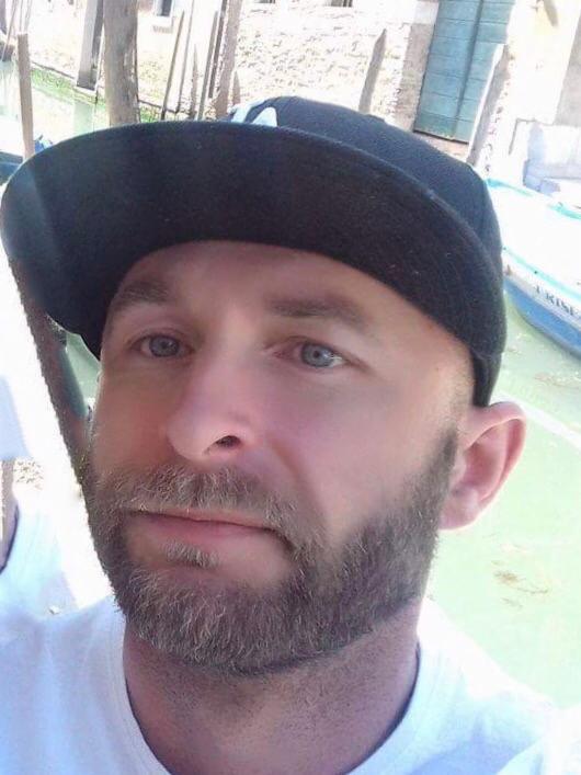 Portret mężczyzny w czapce z lekkim zarostem na tle kanału z łodzią w Wenecji.