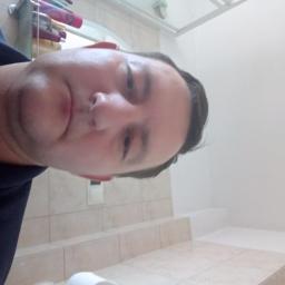 Grumi - Autoportret mężczyzny w łazience z beżowymi płytkami, białą ścianą i sufitem. Widoczne kosmetyki na półce. Nietypowy kąt ujęcia.