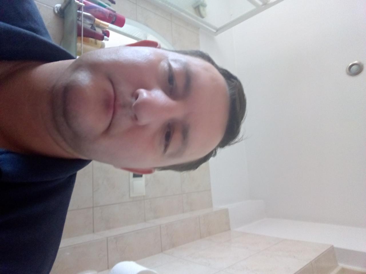 Autoportret mężczyzny w łazience z beżowymi płytkami, białą ścianą i sufitem. Widoczne kosmetyki na półce. Nietypowy kąt ujęcia.