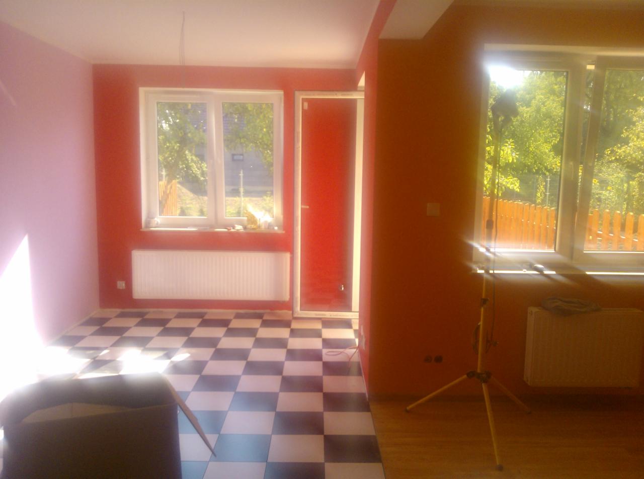 Pokój po malowaniu ścian na odcienie czerwieni i różu, z podłogą w czarno-białą szachownicę, oświetlony lampą budowlaną. Widoczne okna z widokiem na zewnątrz.