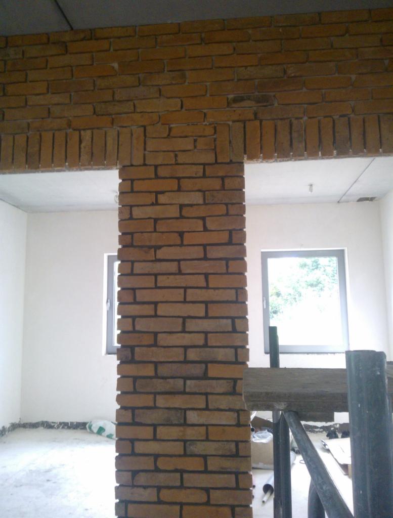 Wnętrze w trakcie wykończenia z kolumną i nadprożem obłożonymi cegłą klinkierową, widoczne rusztowanie i okno z zielenią za nim.