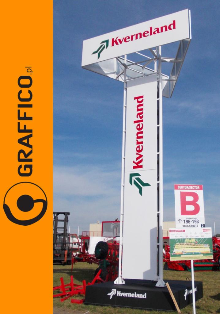 Wysoki, biały pylon reklamowy z logo Kverneland na tle błękitnego nieba, widoczny na tle sprzęt rolniczy i pomarańczowy baner z logotypem GRAFFICO.pl