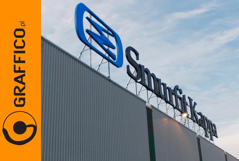 Instalacja przestrzennego logo firmy Smurfit Kappa na dachu hali produkcyjnej, widok z dołu, niebieski element graficzny i czarny napis na tle błękitnego nieba.