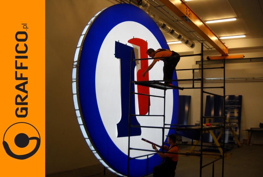 Montaż podświetlanego, okrągłego szyldu z logo w kolorach niebieskim, białym i czerwonym; dwóch pracowników na rusztowaniu podczas instalacji.