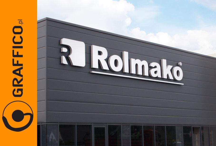 Elewacja budynku z logo firmy Rolmako wykonanym z białych, przestrzennych liter, widoczne z dołu pod lekkim kątem. W lewym dolnym rogu logo firmy GRAFFICO.pl na pomarańczowym tle.