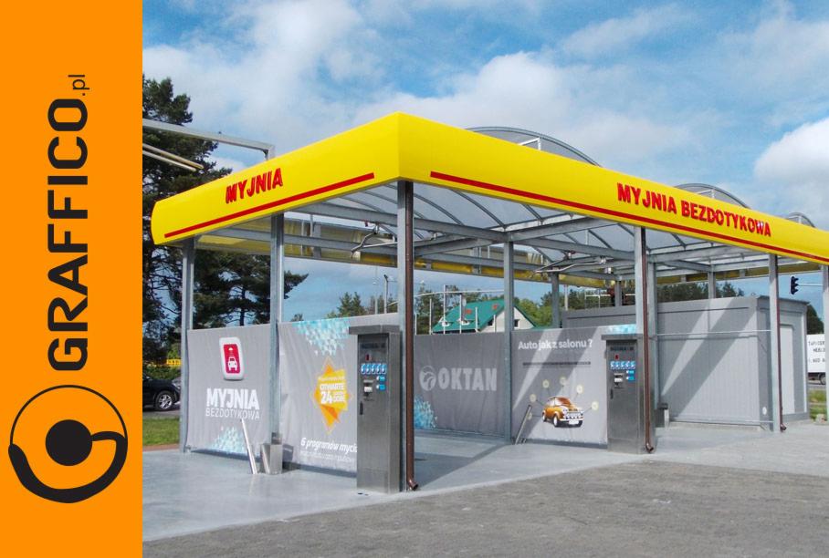 Zewnętrzny widok bezdotykowej myjni samochodowej z żółtym zadaszeniem i szarymi ścianami, automatami płatniczymi i reklamami Oktan, otwartej 24h, z logo firmy Graffico.pl po lewej stronie.