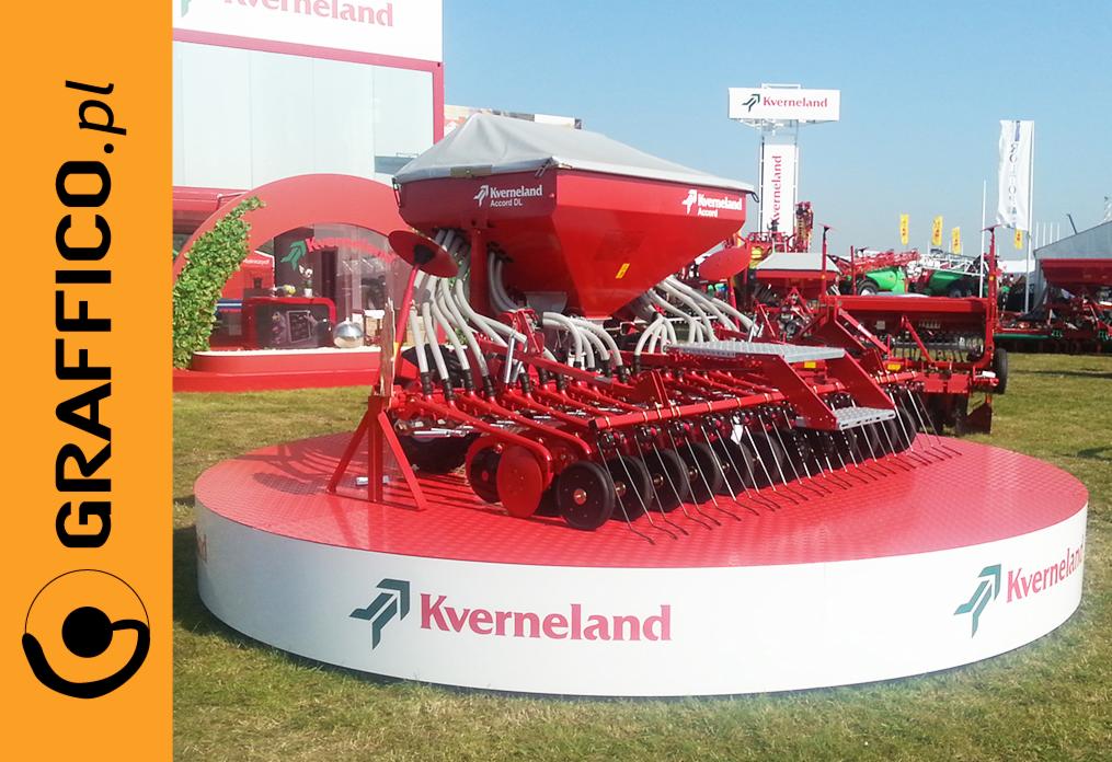 Czerwony siewnik Kverneland na białym podeście z logo, prezentowany na targach rolniczych, z innymi maszynami w tle.