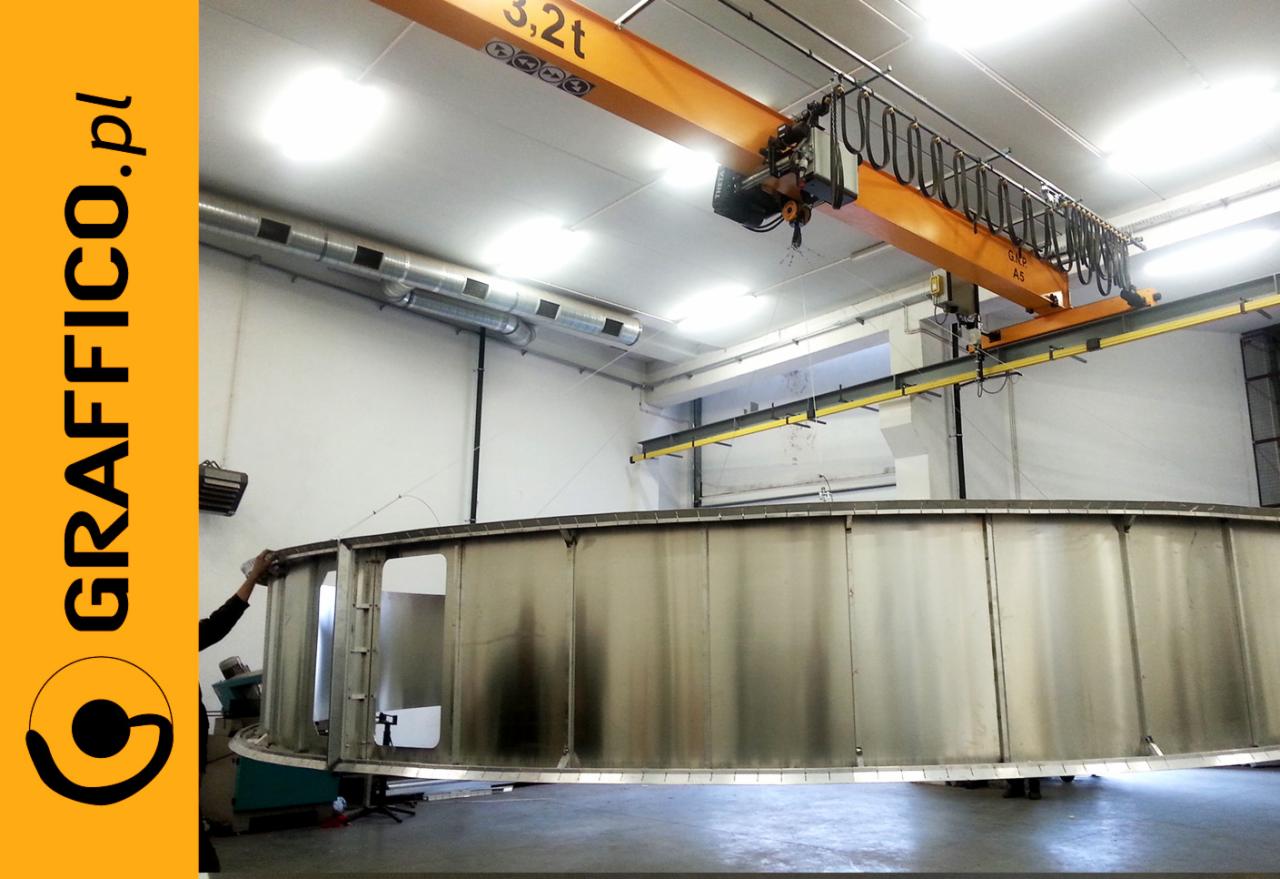 Wnętrze hali produkcyjnej w Toruniu z suwnicą podnoszącą duży, metalowy element konstrukcyjny o okrągłym kształcie, prawdopodobnie w trakcie montażu lub obróbki.