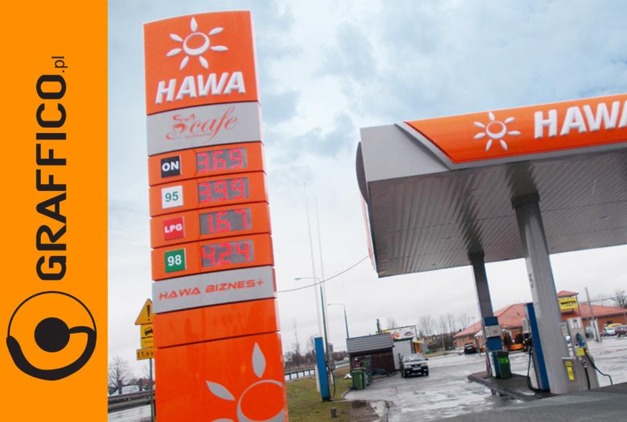 Pylon cenowy stacji paliw Hawa z wyświetlanymi cenami paliw, widoczny dystrybutor, logo firmy na pylonie i zadaszeniu, pochmurne niebo w tle.