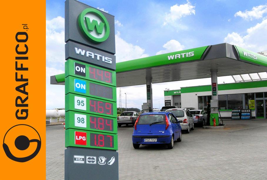 Pylon cenowy stacji benzynowej Watis z aktualnymi cenami paliw, widok na budynek stacji i zaparkowane samochody w tle.