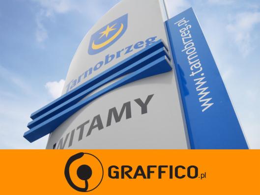 Pylon reklamowy z logo i nazwą Tarnobrzeg oraz adresem strony internetowej, widoczne trzy niebieskie pasy dekoracyjne, błękitne niebo w tle.