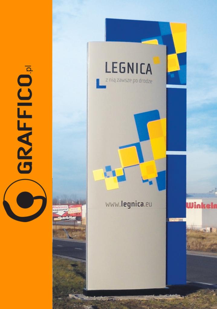 Pylon reklamowy z logo miasta Legnica, z geometrycznym wzorem w kolorach niebieskim i żółtym, umieszczony przy drodze w pobliżu budynków handlowych.
