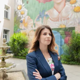 Kobieta w granatowej marynarce, stojąca na dziedzińcu z fontanną, z muralem przedstawiającym postać z różowymi włosami w tle.