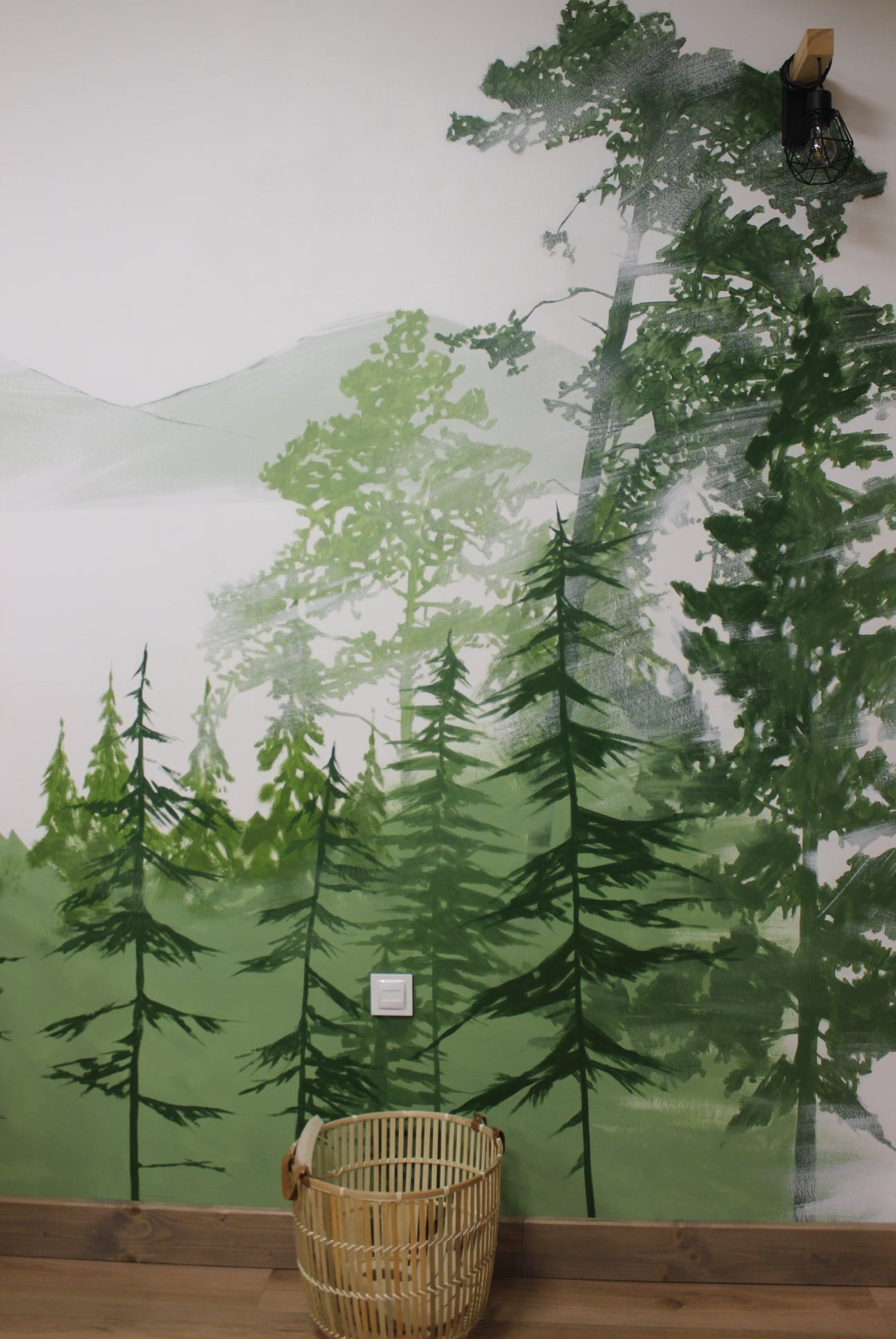 Ściana z ręcznie malowanym pejzażem leśnym w odcieniach zieleni, widoczne drzewa iglaste i liściaste, góry w tle, lampa dekoracyjna na ścianie i wiklinowy kosz na podłodze.