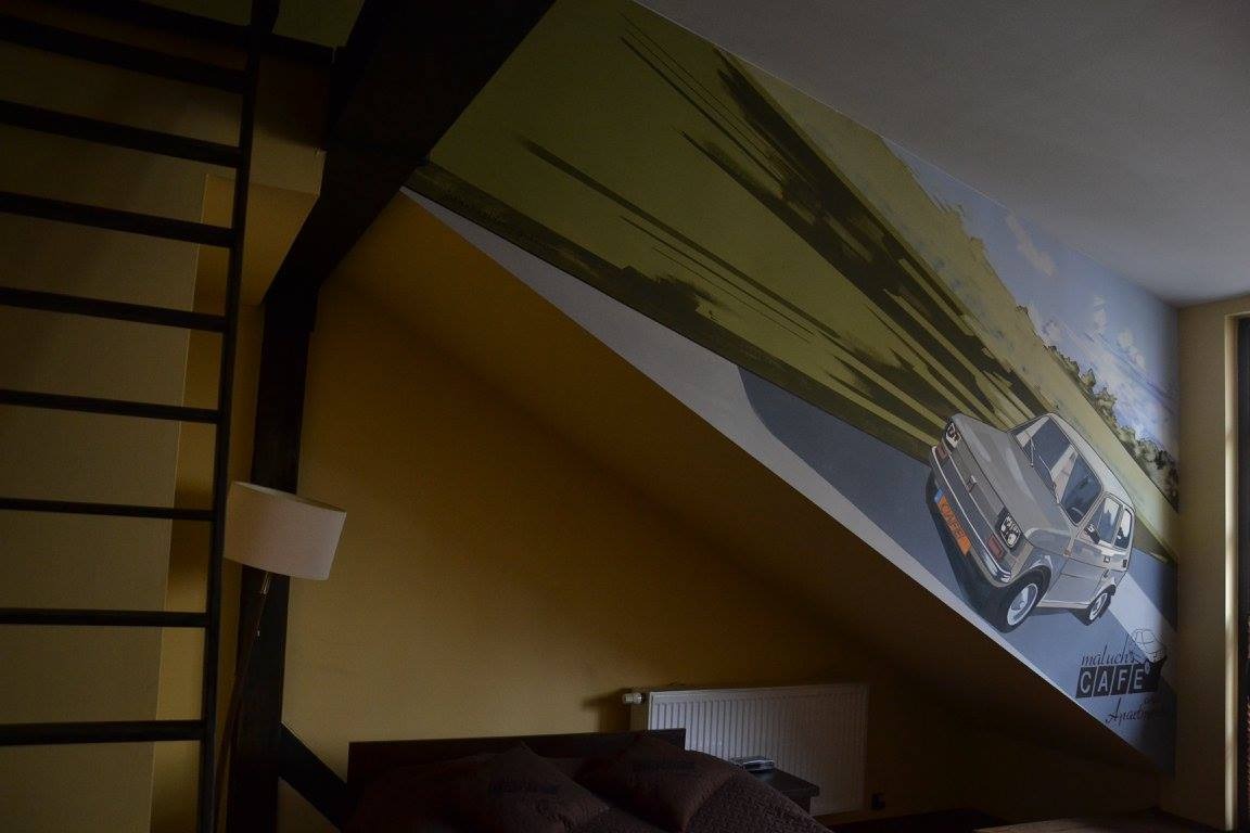 Pokój na poddaszu z muralem przedstawiającym szarego Fiata 126p na drodze, z napisem 'maluch CAFE', drabina i lampa widoczne po lewej stronie.