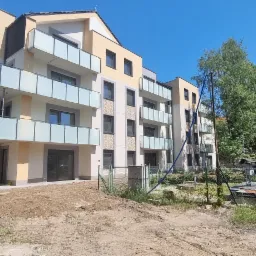 Nowoczesny budynek wielorodzinny z balkonami o szklanych balustradach, zlokalizowany w sąsiedztwie zieleni, widoczny teren przygotowany pod zagospodarowanie.