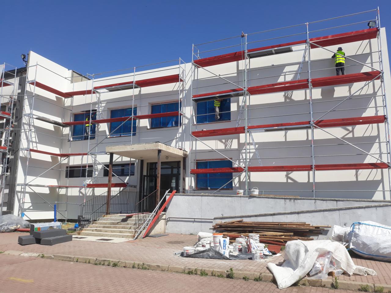 Budynek w trakcie prac elewacyjnych z użyciem rusztowań, okna zabezpieczone folią, widoczne puszki z farbą i materiały budowlane na chodniku.