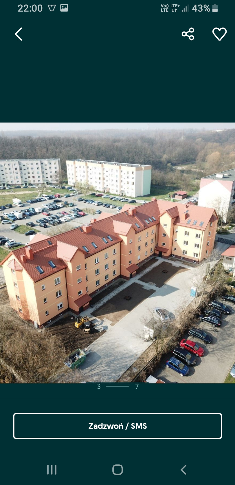Widok z góry na nowo wybudowany, pomarańczowy budynek wielorodzinny z czerwonym dachem, z placem budowy, małą koparką i kontenerem na śmieci przed budynkiem, na tle bloków mieszkalnych i drzew.