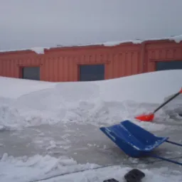 Dach budynku zasypany śniegiem, częściowo odśnieżony, widoczne dwie pomarańczowe i jedna niebieska łopata do śniegu