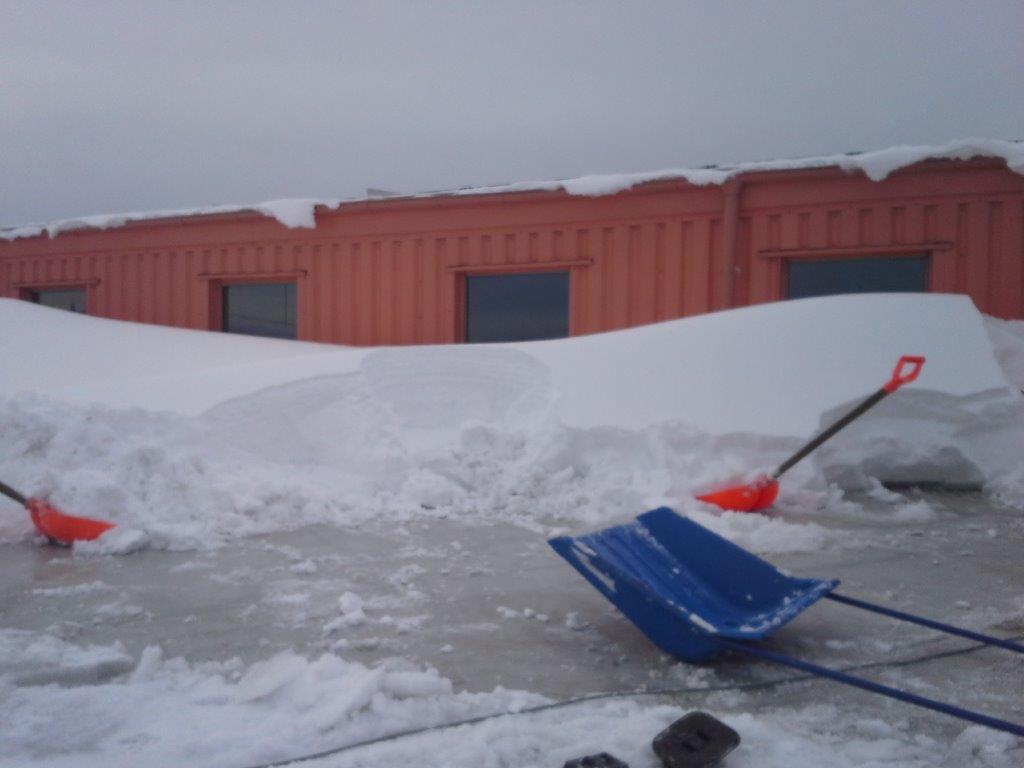 Dach budynku zasypany śniegiem, częściowo odśnieżony, widoczne dwie pomarańczowe i jedna niebieska łopata do śniegu