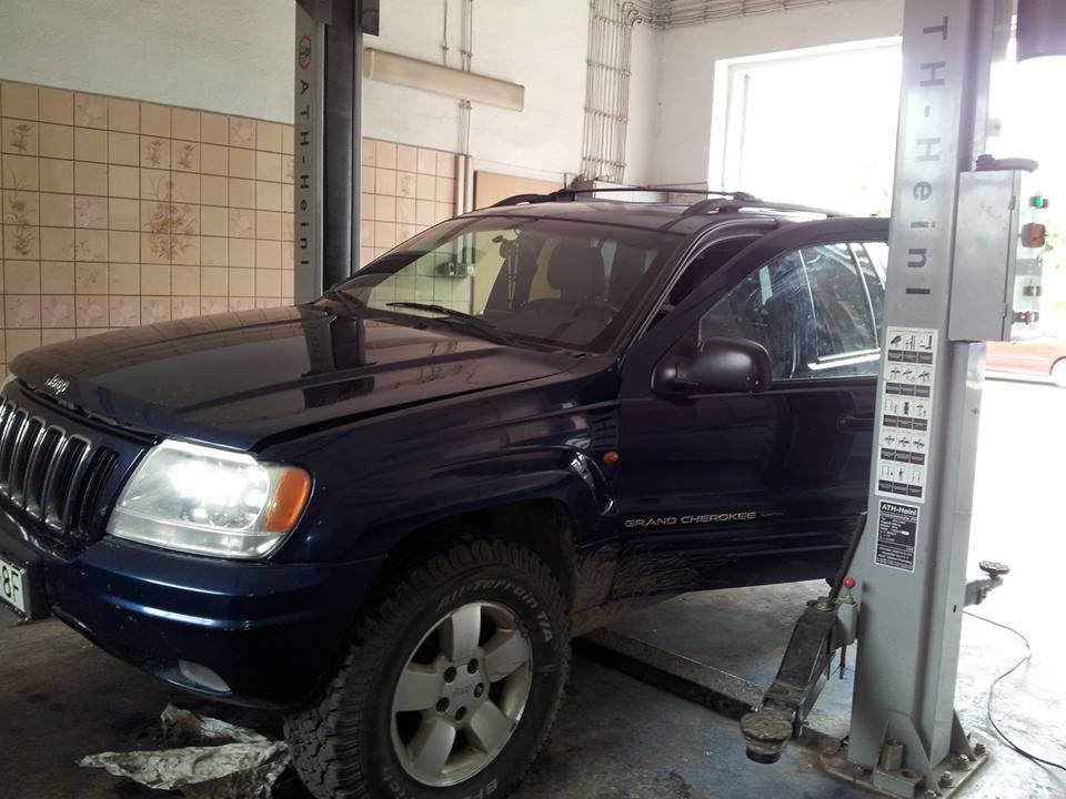 Niebieski Jeep Grand Cherokee podniesiony na podnośniku samochodowym w warsztacie, widoczne otwarte drzwi kierowcy i opony terenowe.