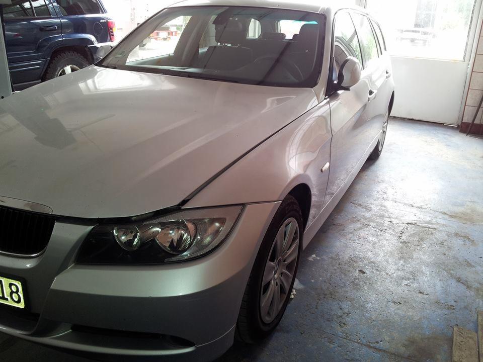 Srebrny samochód kombi z uszkodzonym przodem, zaparkowany w garażu z betonową posadzką, widoczny fragment niebieskiego SUV-a w tle.