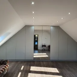 Garderoba na poddaszu ze szklanymi drzwiami, szare fronty, drewniana podłoga, widoczna sofa z pledem w kratę.